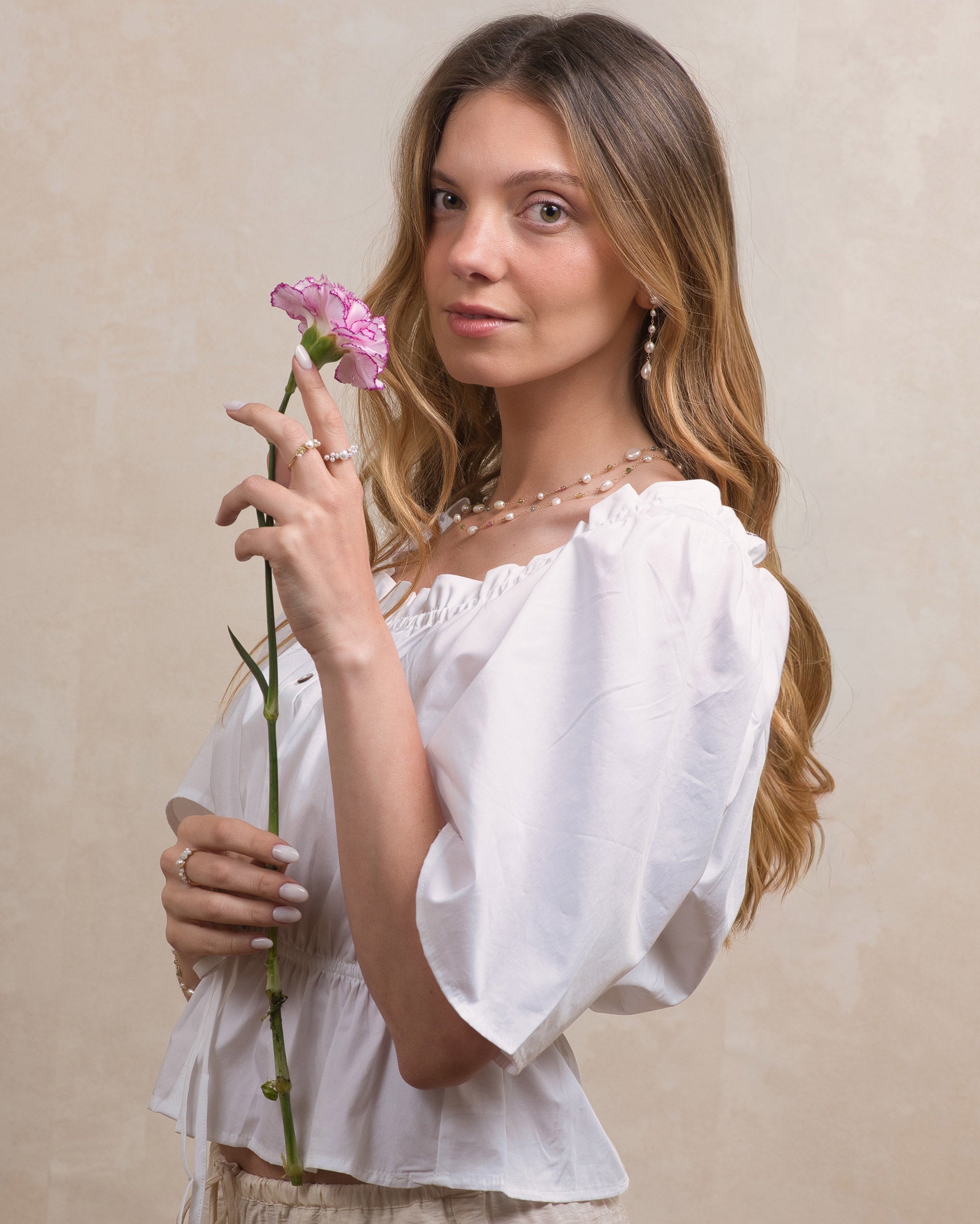Woman in a white blouse holding a pink flower, wearing a Mamie Coco keshi pearl and tourmaline double row necklace and Mamie Coco long pearl earrings, against a beige background