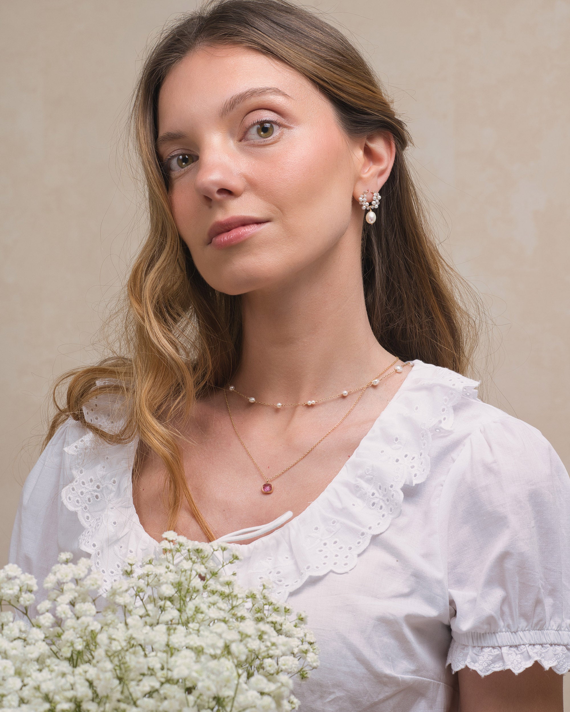Woman wearing a white blouse with lace details, holding flowers, and wearing Mamie Coco bridal pearl earrings, a pearl necklace layered with a gold bezel pink tourmaline pendant, against a beige background.