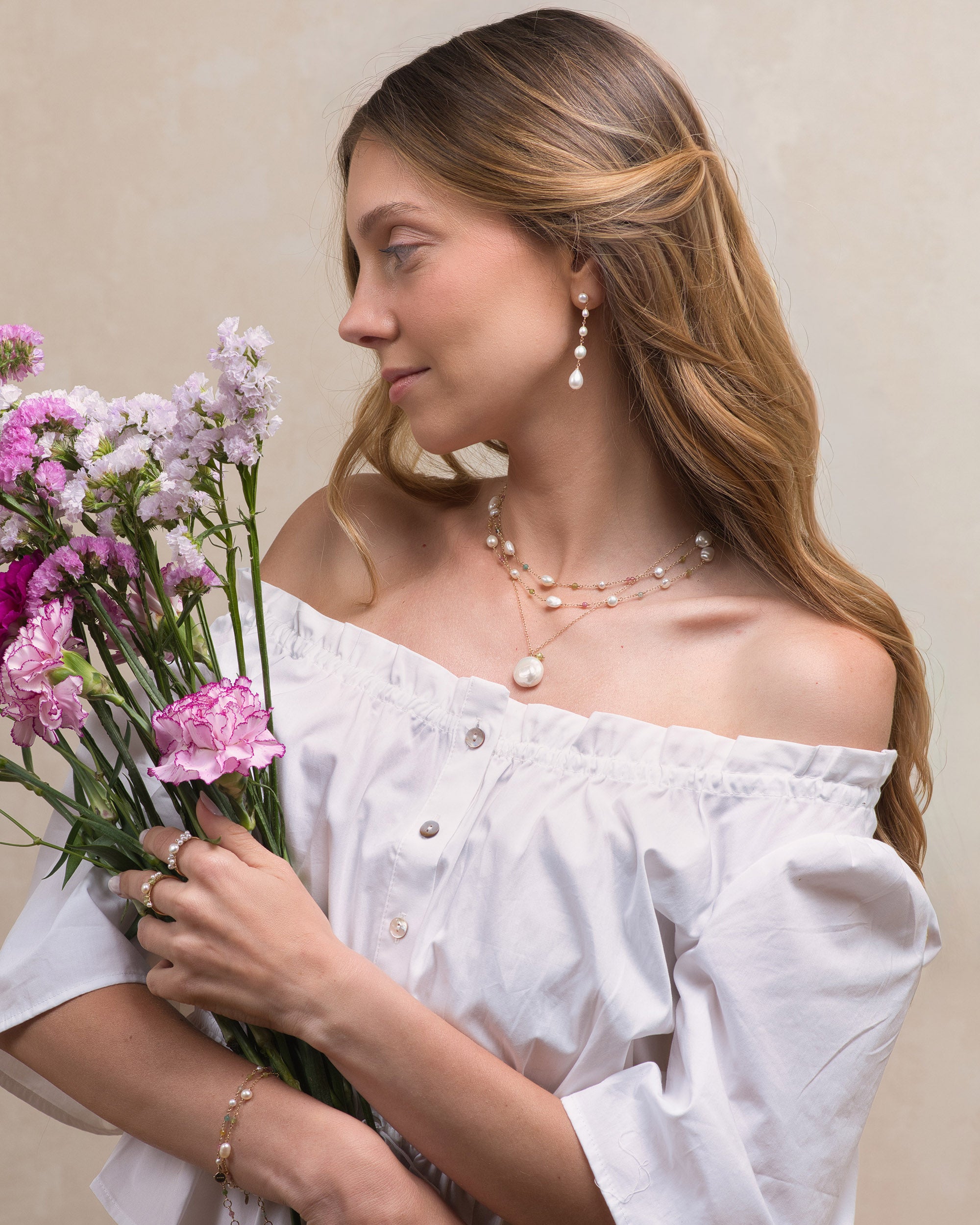 Woman holding flowers and wearing  a Mamie Coco keshi pearl and tourmaline double row necklace  layered with a large baroque pearl pendant and long pearl earrings, against a neutral background