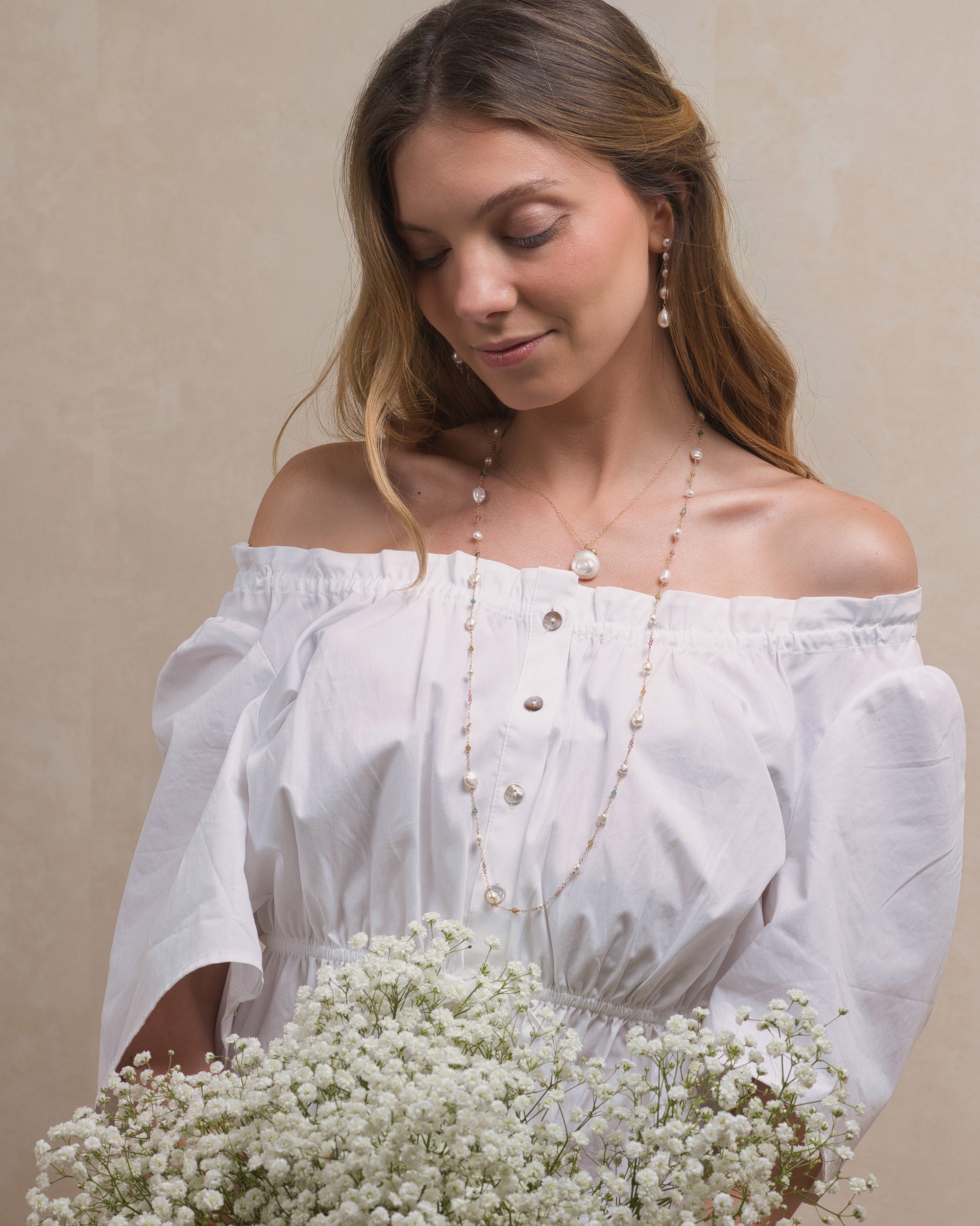 Woman in a white off-shoulder blouse holding a bouquet of white flowers and wearing a long Mamie Coco keshi pearl and tourmaline necklace and long pearl earrings, against a beige background